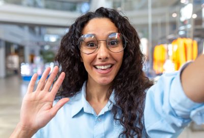 A young beautiful happy Latin American woman is standing in a shopping center, talking on a video call, waving, greeting, smiling at the camera.