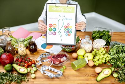 Dietitian showing a scheme with vitamins and minerals sitting at the table fulll of variuos healthy products indoors. Healthy eating concept